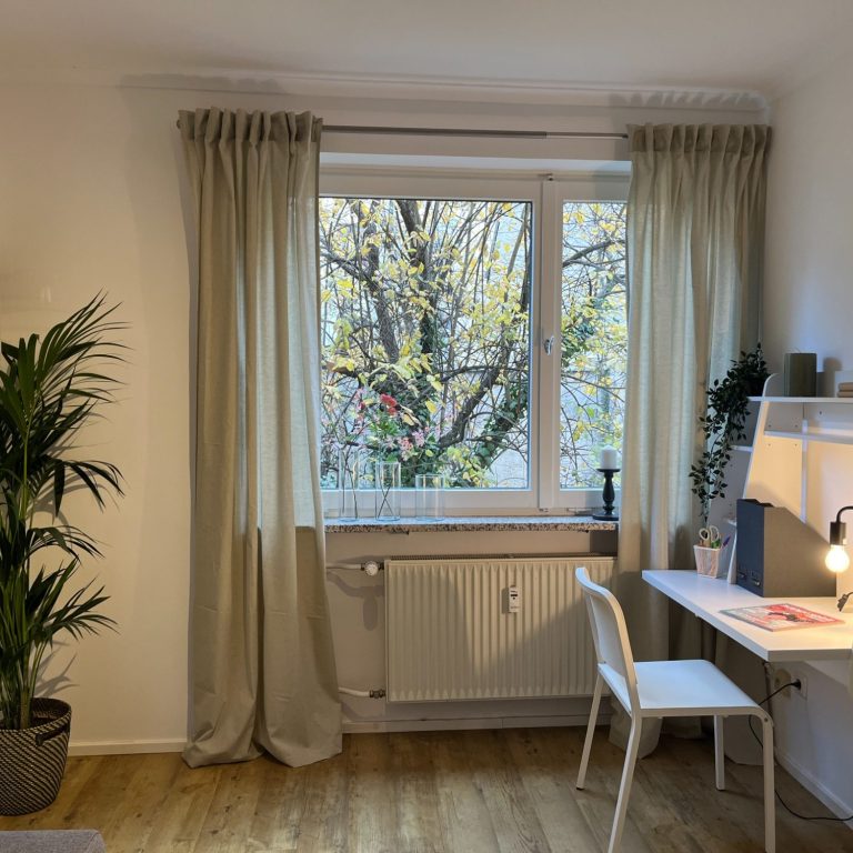 Wohnzimmer_nachher Heller gemütlicher Raum, dekoriert mit Vorhängen, mit einem Schreibtisch, einer großen Pflanze und einem Fenster mit Blick auf Bäume.