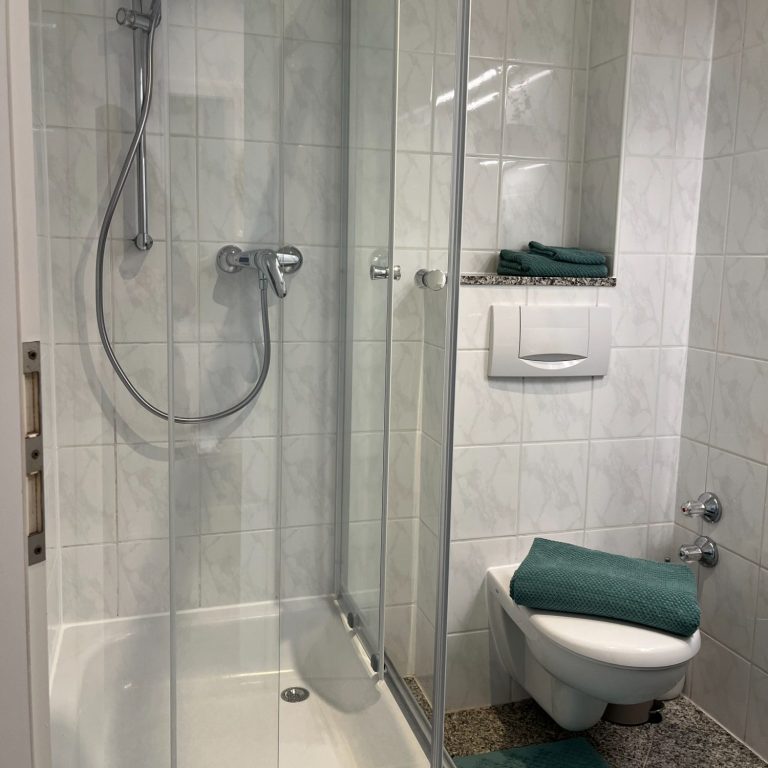 Bad_nachher saubere, neue Dusche mit Glaswand, WC und türkisfarbene Handtücher in einem hellen Badezimmer.