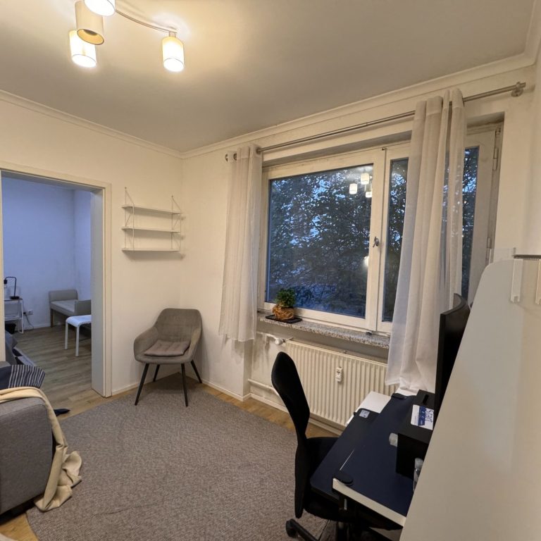 Wohnzimmer_vorher Heller Raum mit Schreibtisch, Stuhl und einem Fenster mit Blick nach draußen.