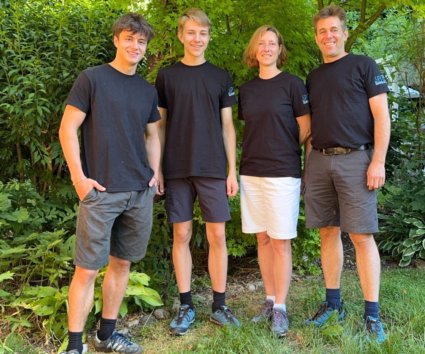 Das Team das Team: Vier Personen in schwarzen T-Shirts stehen in einem Garten mit grüner Umgebung.