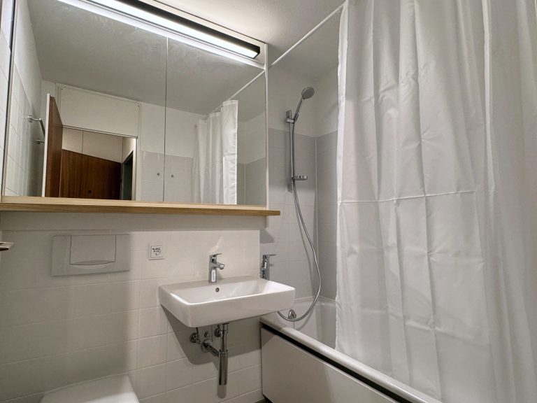 Spiegelschrank mit Holzablage Kleines Badezimmer mit Dusche, Waschtisch und Spiegel mit Holz-Ablage, helle Wände und Vorhang.